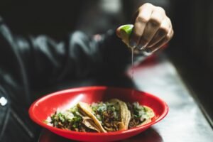 Close-up of a hand squeezing lime over tasty Mexican street tacos in a red plate, highlighting fresh ingredients.